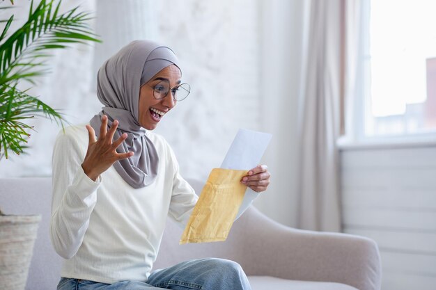 A young muslim woman in a hijab is sitting on the sofa at home and reading a received letter in an