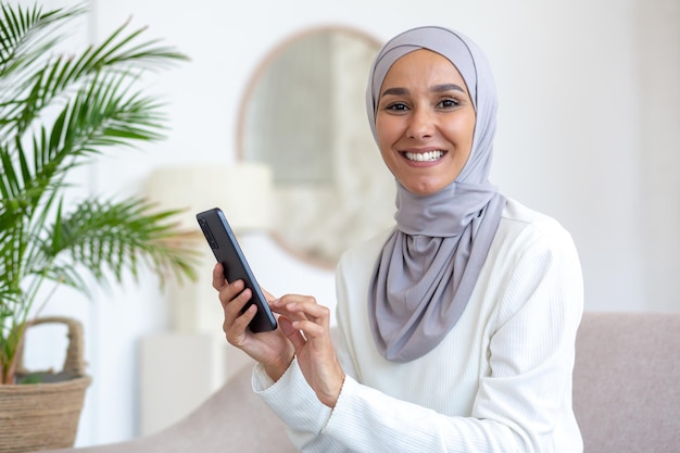 Young muslim woman in hijab sitting on sofa at home and using phone smiling she looks at the camera