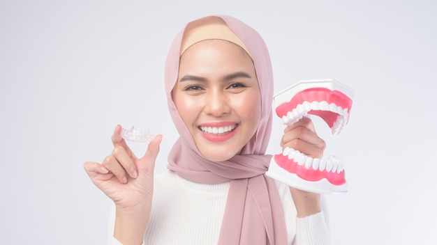 Молодая мусульманка держит подтяжки invisalign и искусственную стоматологическую модель человека на белом, стоматологическом здравоохранении и ортодонтической концепции.