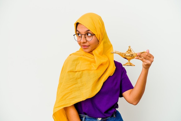 Young muslim woman holding a lamp isolated on white background looks aside smiling, cheerful and pleasant.