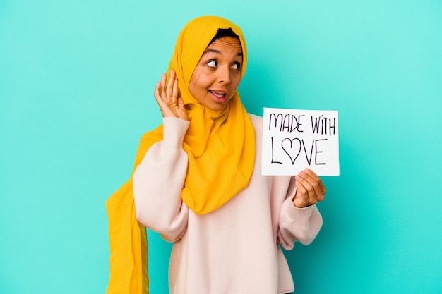 Young muslim woman holding a made with love placard on blue trying to listening a gossip.