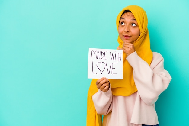 Young muslim woman holding a made with love placard isolated on blue background looking sideways with doubtful and skeptical expression.