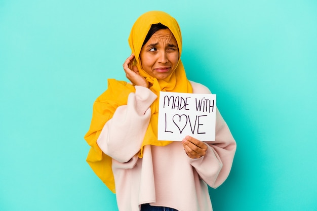 Young muslim woman holding a made with love placard isolated on blue wall covering ears with hands.