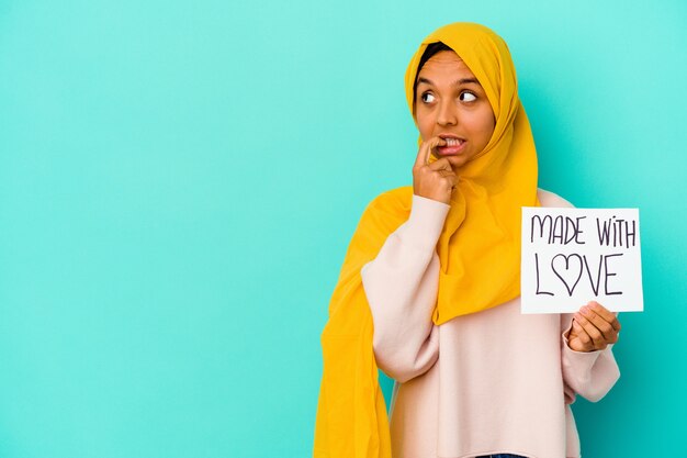 Young muslim woman holding a made with love placard isolated on blue wall relaxed thinking about something looking at a copy space.