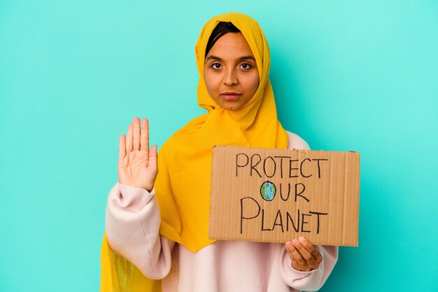 Young muslim woman holding a protect our planet isolated on blue background standing with outstretched hand showing stop sign, preventing you.