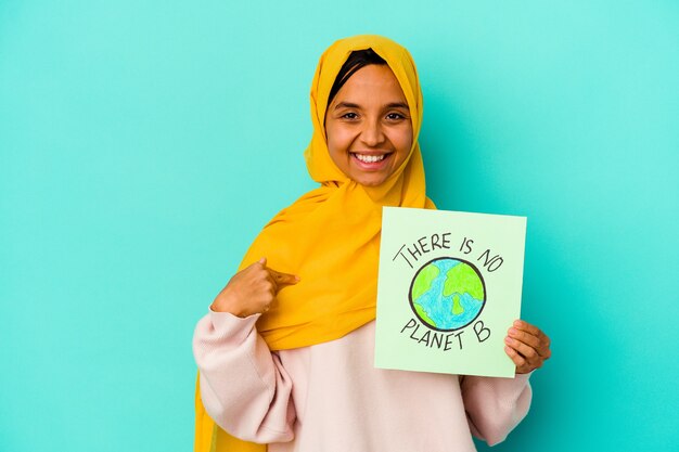 Young muslim woman holding a there is not planet b placard  isolated on blue background person pointing by hand to a shirt copy space, proud and confident