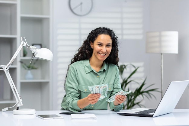 A young muslim woman is sitting in the office at a desk she holds cash in her hands received a