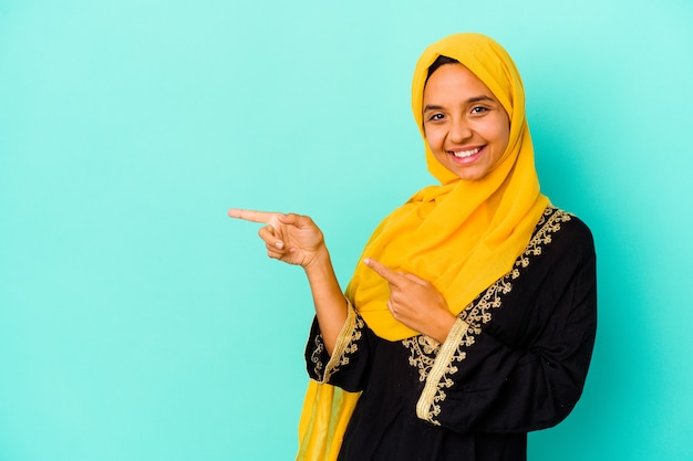 Young muslim woman isolated on blue background excited pointing with forefingers away.