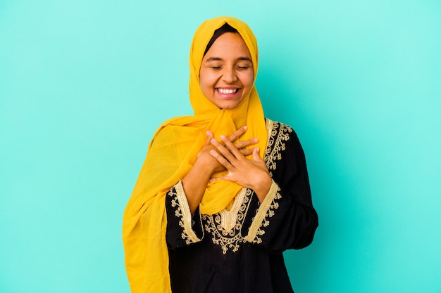 Young muslim woman isolated on blue background laughs out loudly keeping hand on chest.