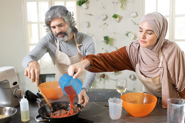 成熟したひげを生やした男性がペストリーの詰め物を調理するのを助けながら、フライパンでミンチ肉にトマトケチャップを注ぐ若いイスラム教徒の女性