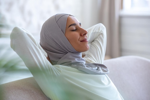 Young muslim woman resting at home with eyes closed hands behind head napping sitting on sofa woman