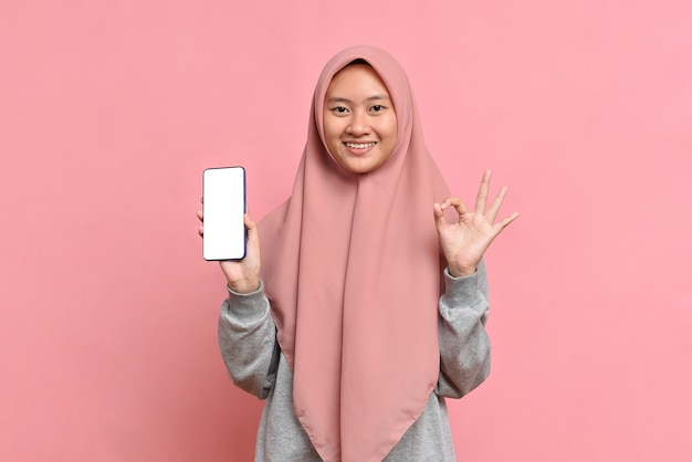 Young Muslim woman showing phone screen display with okay sign against on pink background