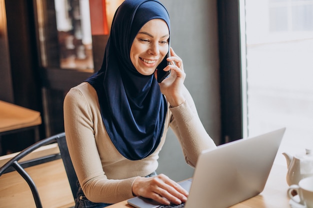 電話を使い、カフェでコンピューターを操作する若いイスラム教徒の女性