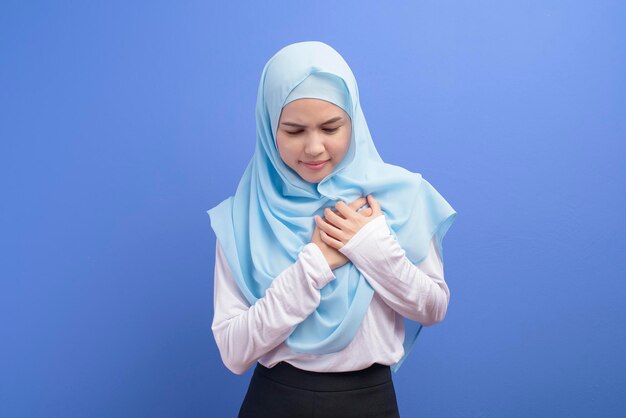 Una giovane donna musulmana che indossa l'hijab che soffre di dolore toracico su sfondo blu studio, attacco cardiaco e concetto medico.