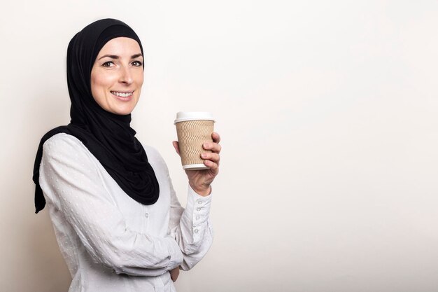 Young muslim woman in a white shirt and hijab with a smile holds a paper cup with coffee banner