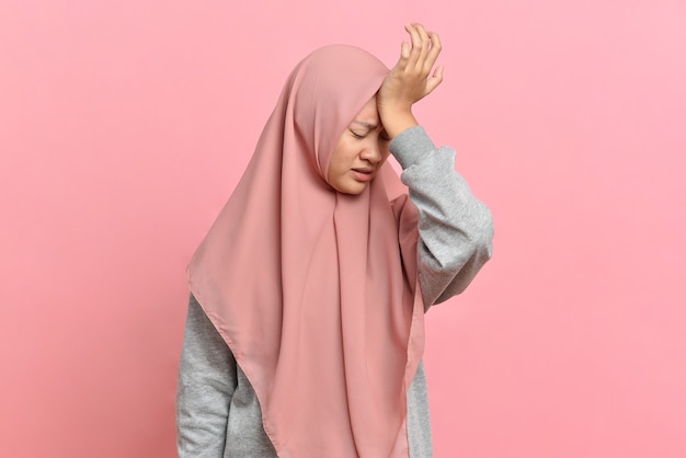 Young muslim woman with a headache holds her temples with her hand. Health, migraine and headache concept, isolated on pink background