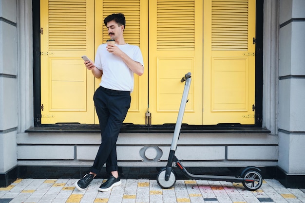 Young mustache man standing with coffee to go and e-scooter while dreamily using cellphone on city street