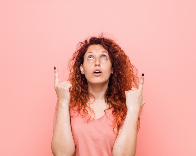 Young natural and authentic redhead woman pointing upside with opened mouth.