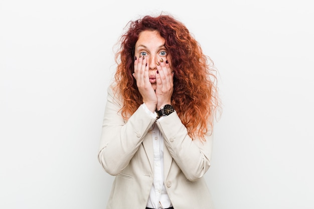 Young natural redhead business woman against white wall blink through fingers frightened and nervous.