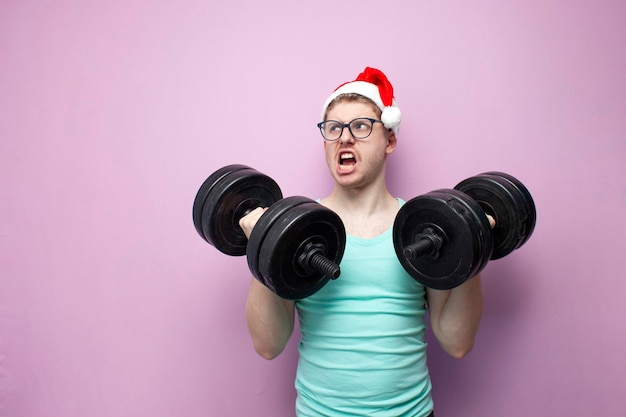 Photo young nerd guy in glasses and santa hat trains and lifts heavy dumbbells on pink background