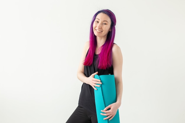 Young nice girl hipster with colored hair holding in hand a sports mat posing on a white wall. Healthy lifestyle concept. 