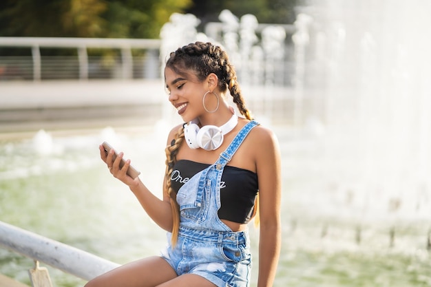 Young and nice girl sits near the fountain in headphones listens to music and looks into telephone