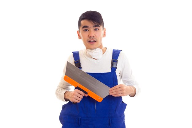 Young nice man with dark hair in white shirt and blue overall with white respirator holding spatula on white background in studio