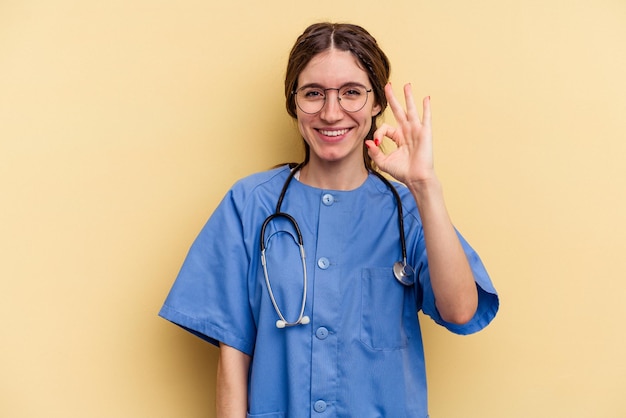 Young nurse caucasian woman isolated on yellow background cheerful and confident showing ok gesture