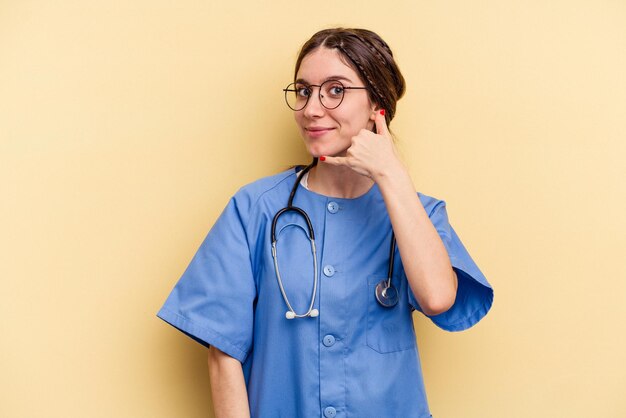 Young nurse caucasian woman isolated on yellow background showing a mobile phone call gesture with fingers.