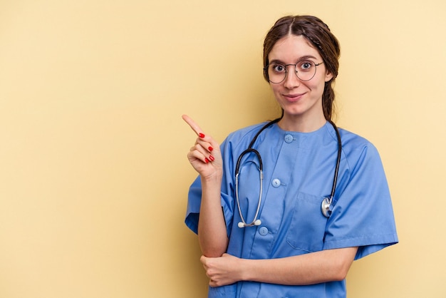 Young nurse caucasian woman isolated on yellow background smiling and pointing aside, showing something at blank space.