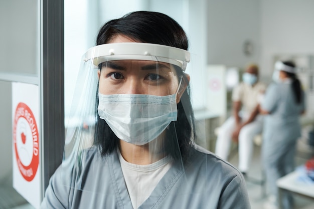 Young nurse or clinician in protective workwear standing in front of camera against her colleague vaccinating male patient in medical office