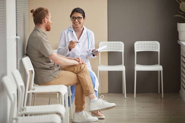 Молодая медсестра в очках улыбается и разговаривает с пациентом, сидя на стульях в коридоре больницы