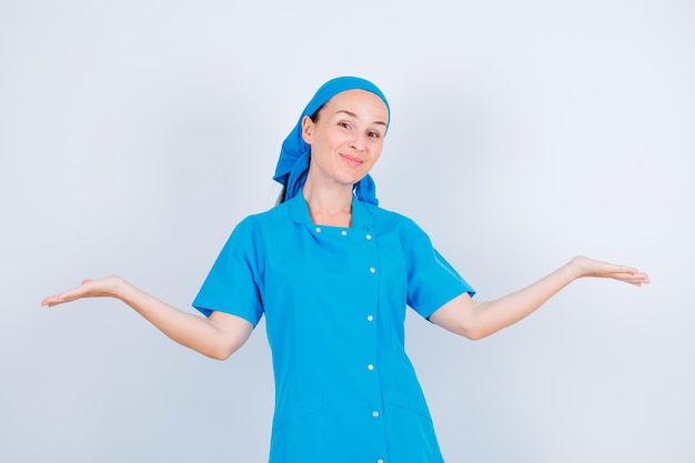 Young nurse is looking at camera by raising up and opening wide her hands on white background