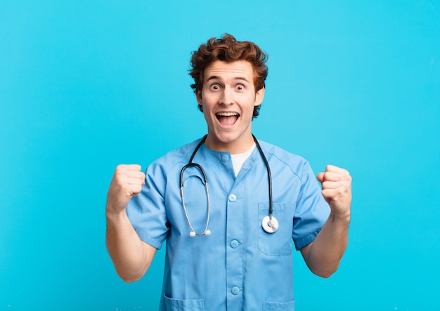 Young nurse man feeling shocked, excited and happy, laughing and celebrating success, saying wow!