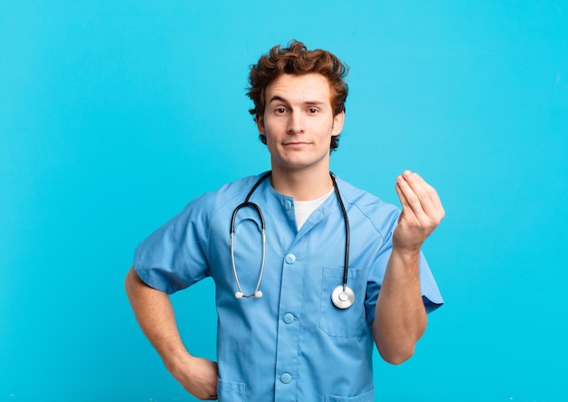 Young nurse man making capice or money gesture, telling you to pay your debts!