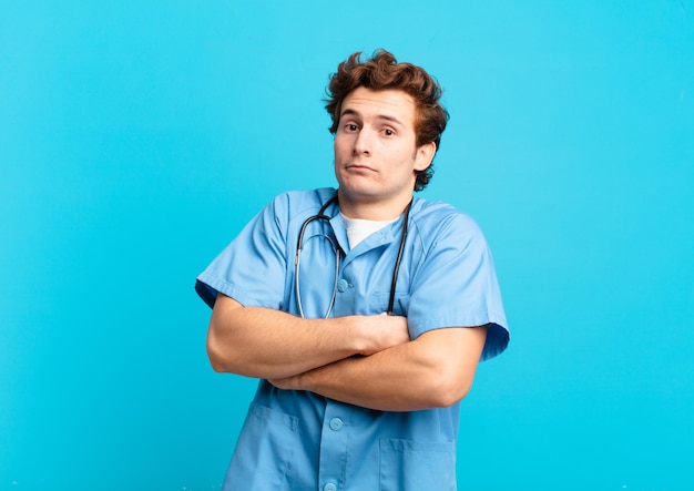 Young nurse man shrugging, feeling confused and uncertain, doubting with arms crossed and puzzled look