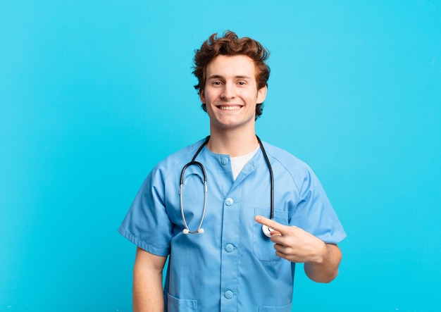 Young nurse man smiling cheerfully feeling happy and pointing to the side and upwards showing object in copy space