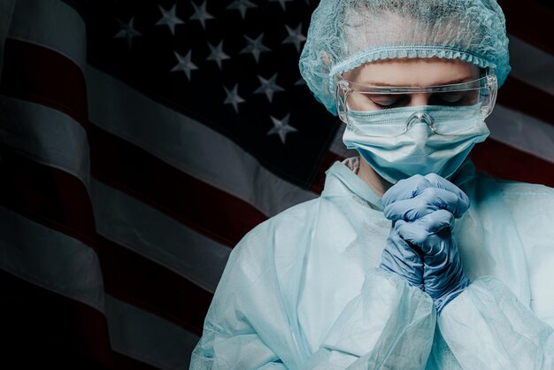 Young nurse in a protective medical mask and hat and dressing gown prays against the backdrop of the American flag and asks God for help and strength in the fight against the coronavirus epidemic