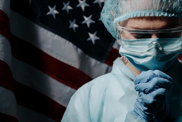 Young nurse in a protective medical mask and hat and dressing gown prays against the backdrop of the American flag and asks God for help and strength in the fight against the coronavirus epidemic