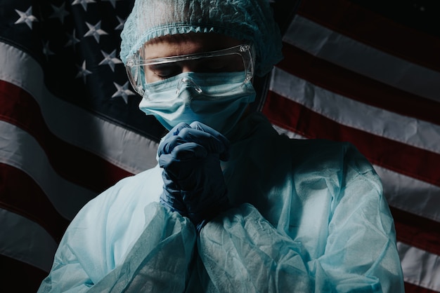 Young nurse in a protective medical mask and hat and dressing gown prays