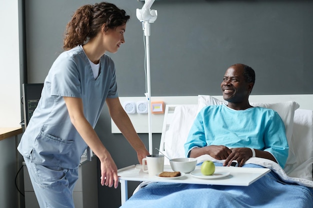 Young nurse in uniform bringing lunch for elderly patient while he sitting on bed in hospital ward