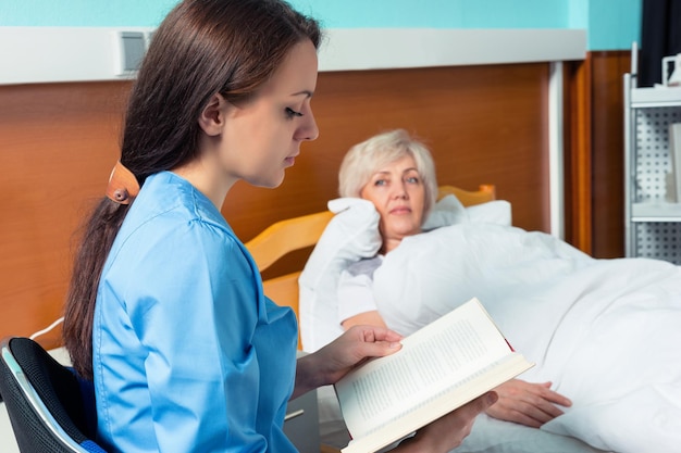 Молодая медсестра в униформе читает книгу и сидит рядом с пациентом, который лежит на больничной койке в палате. Концепция здравоохранения