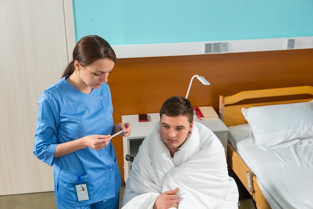 Young nurse in uniform looking at a thermometer of patient, who is sitting on wheelchair covered with quilt in hospital ward. Healthcare concept