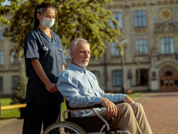 車椅子で成熟した男性患者を押すフェイスシールドとマスクを身に着けている若い看護師