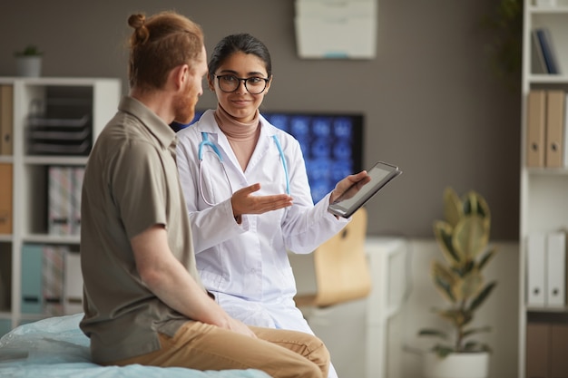 Young nurse in white coat showing medical recipe on digital tablet and talking to the patient during his visit at hospital