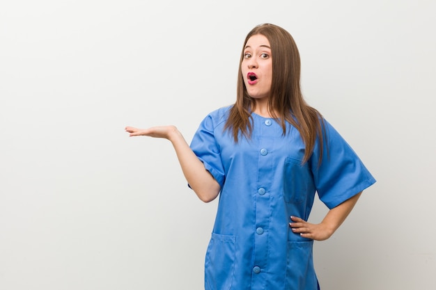 Young nurse woman against a white wall impressed holding copy space on palm.