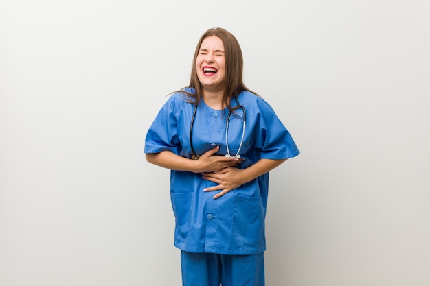 Young nurse woman against a white wall laughs happily and has fun keeping hands on stomach.