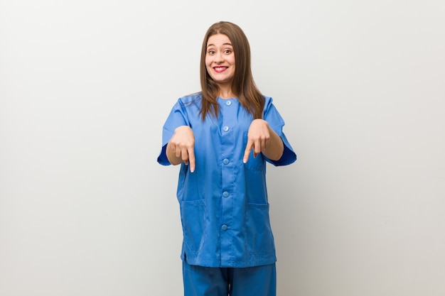 Young nurse woman against a white wall points down with fingers, positive feeling.