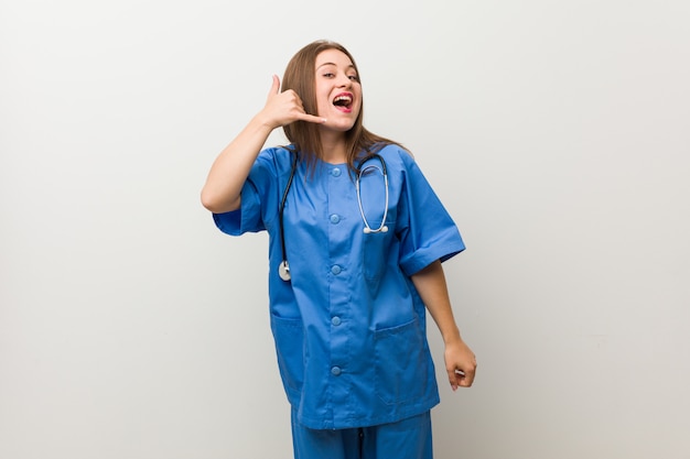 Young nurse woman against a white wall showing a mobile phone call gesture with fingers.