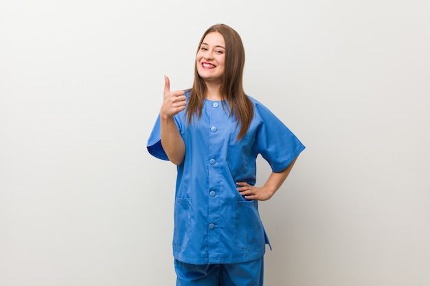 Young nurse woman against a white wall smiling and raising thumb up